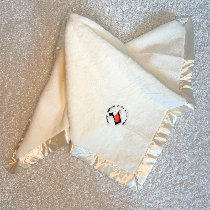 allied blanket - Baby Showers by Mail White plush blanket with a cream satin border, folded on a light carpet, featuring a small circular embroidered logo at the center.