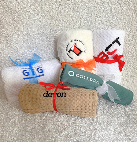 corporate-hp-updated - Baby Showers by Mail Five small rolled towels tied with colorful ribbons (blue, orange, red, white) arranged on a soft, textured surface; each towel displays a different brand logo.