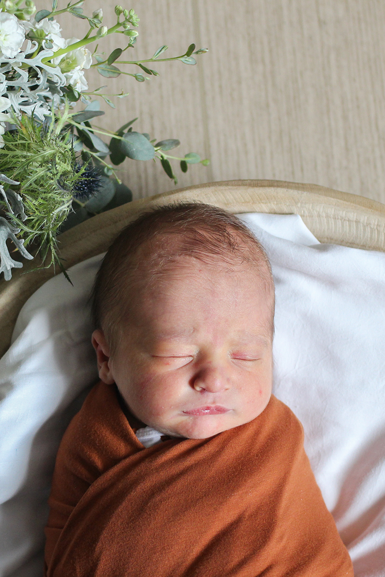 Newborn baby sleeping, wrapped in an orange blanket on a white sheet with a bouquet of greenery and white flowers nearby on the left.