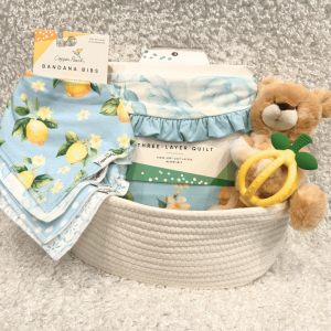 Baby gift basket with lemon-pattern bandana bibs, a blue three-layer quilt, and a plush bear with a yellow teether ring on a soft rug.